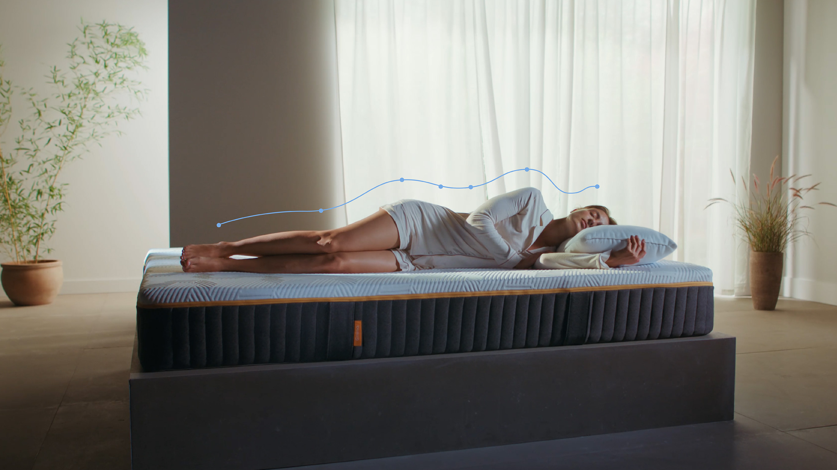 Woman lying on her side on an Emma Original Elite mattress with a spinal alignment graphic overlay showing body support in a bright room.