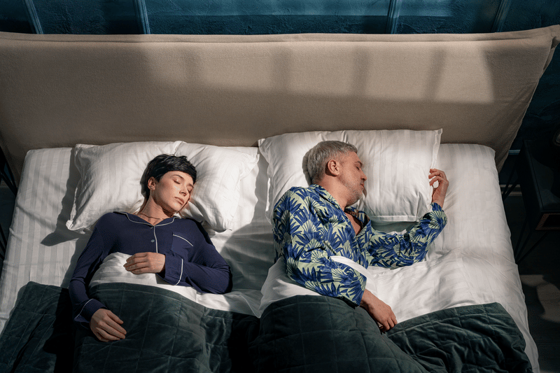 Twee mensen liggen in bed onder donkergroene dekens; links persoon in donkerblauwe pyjama, rechts persoon in blauwgroen patroonshirt; witte lakens, beige hoofdeinde en blauwe muur op de achtergrond.