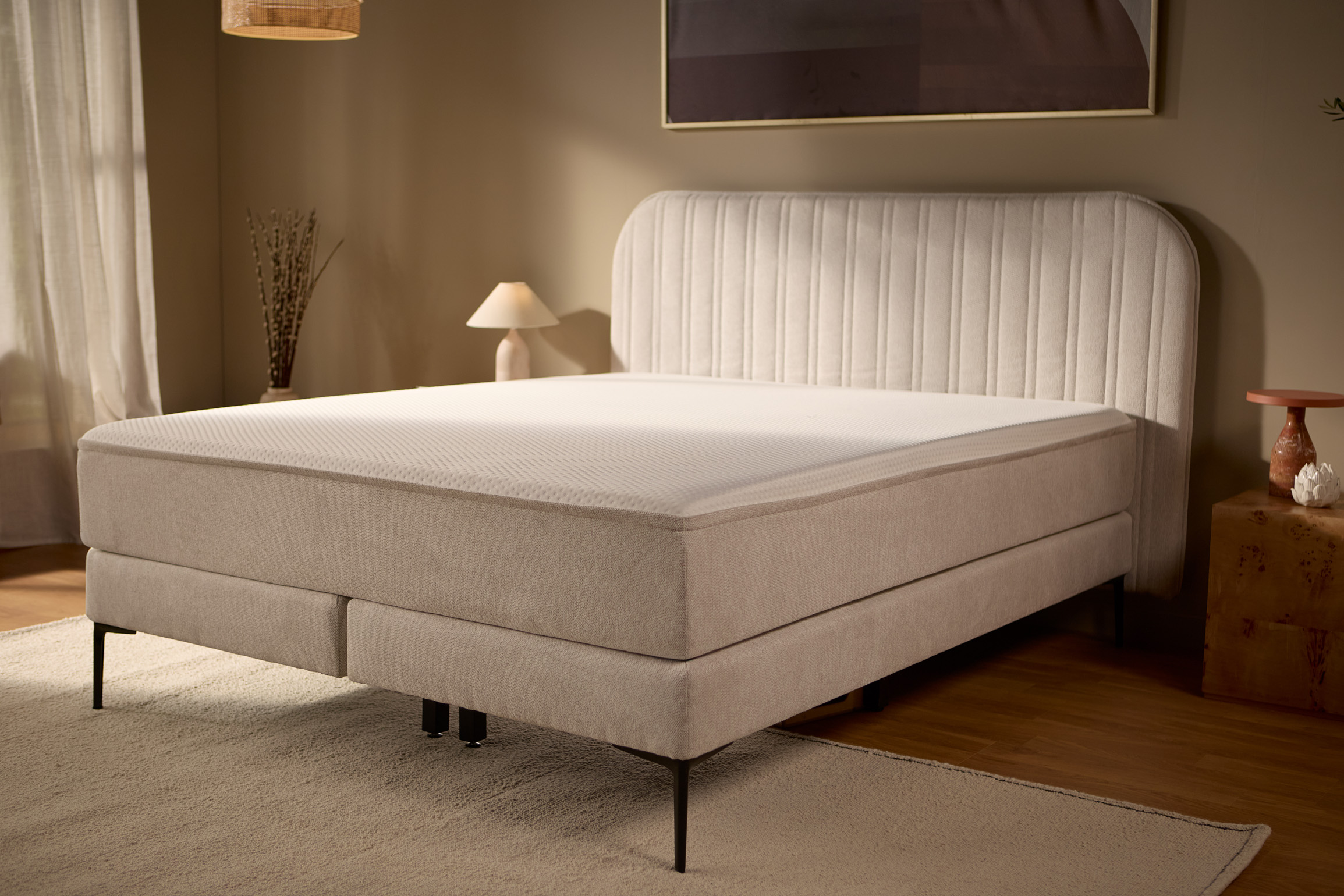 Schuine aanzicht van het bed met de gestoffeerde basis, het hoofdbord met verticale stiknaden en centrale metalen poten voor extra ondersteuning.