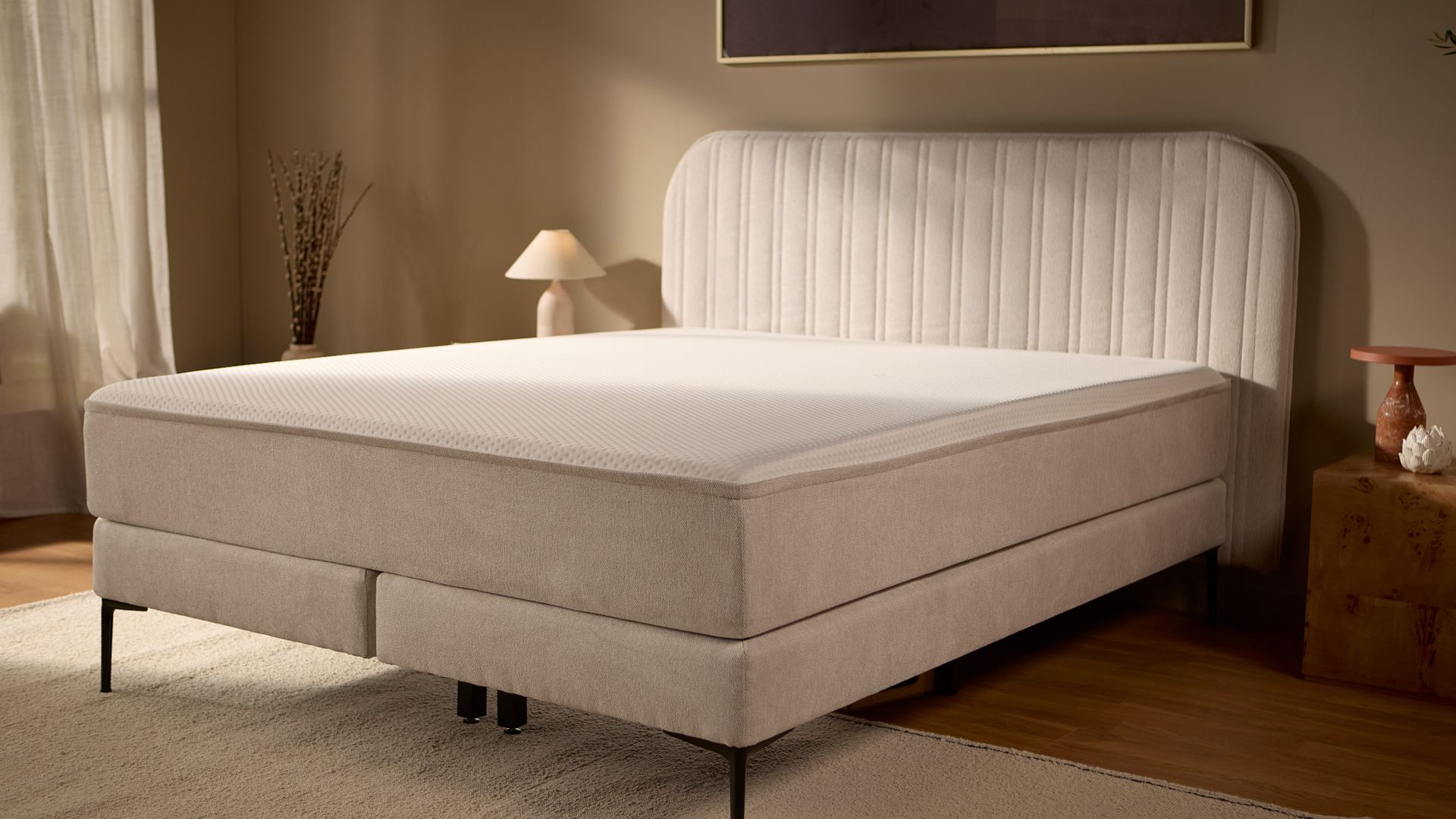 Schrägansicht des Bettes mit gepolstertem Untergestell, Kopfteil mit Kanalsteppung und mittigen Metallfüßen für zusätzlichen Halt.