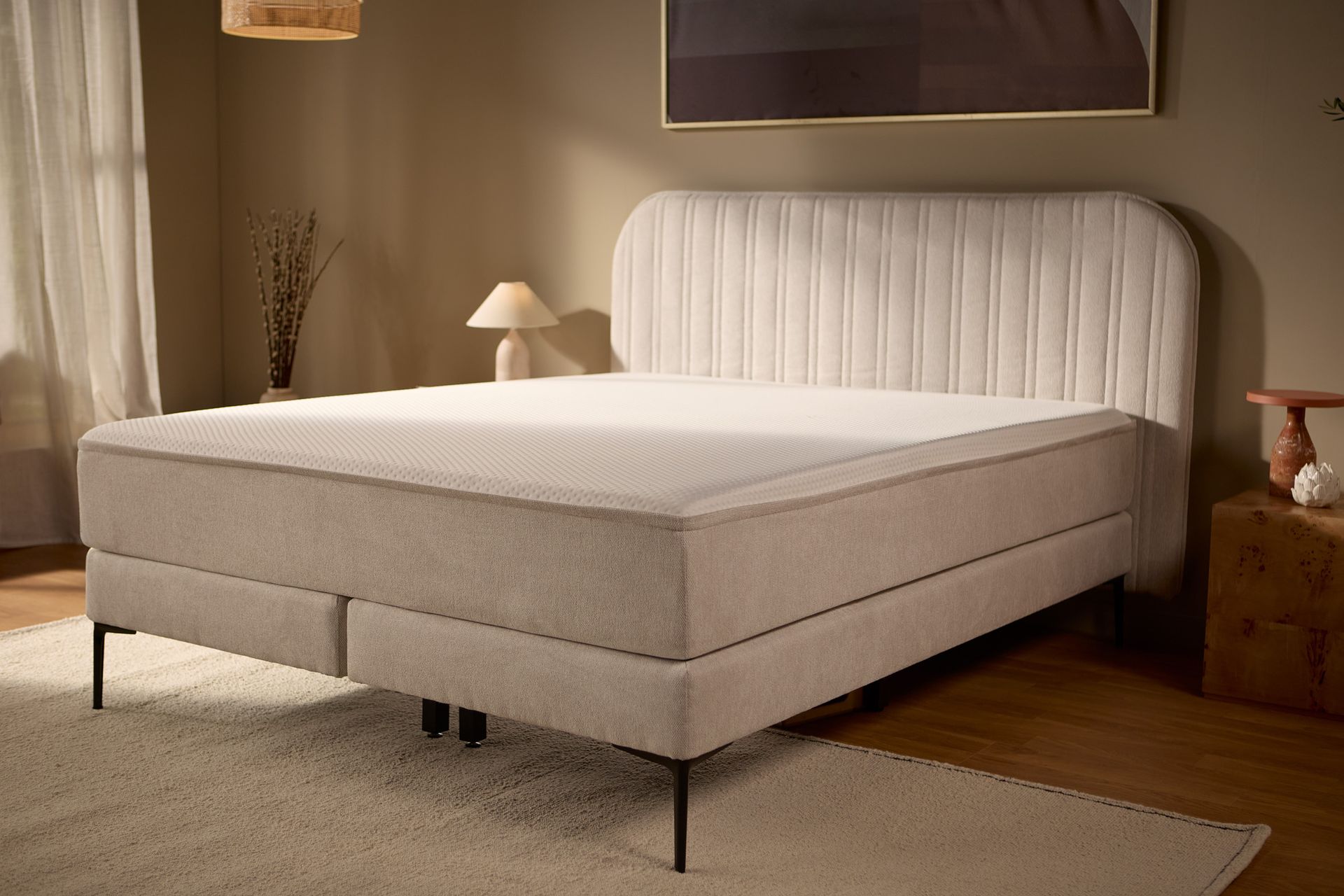 Schuine aanzicht van het bed met de gestoffeerde basis, het hoofdbord met verticale stiknaden en centrale metalen poten voor extra ondersteuning.