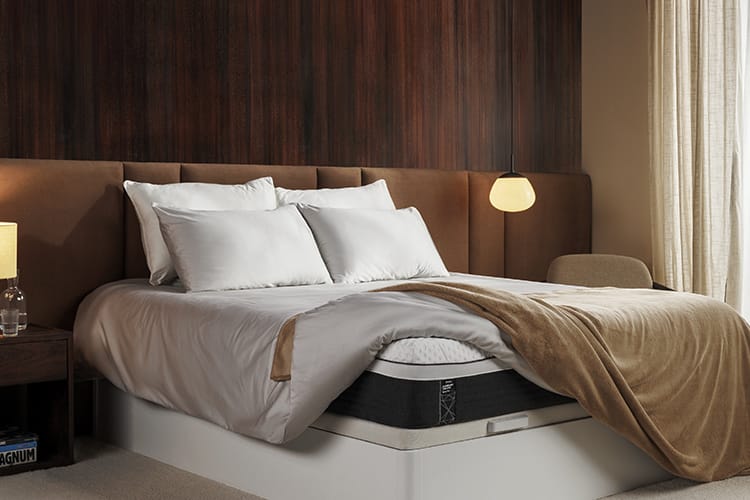 Een modern ingerichte slaapkamer met een opgemaakt bed, witte kussens, een beige plaid en een gestoffeerd bruin hoofdbord. Links staat een houten nachtkastje met een lamp en flesje, rechts hangt een hanglamp boven een stoel. Een houten accentmuur en lichte gordijnen zorgen voor een warme, rustgevende sfeer.