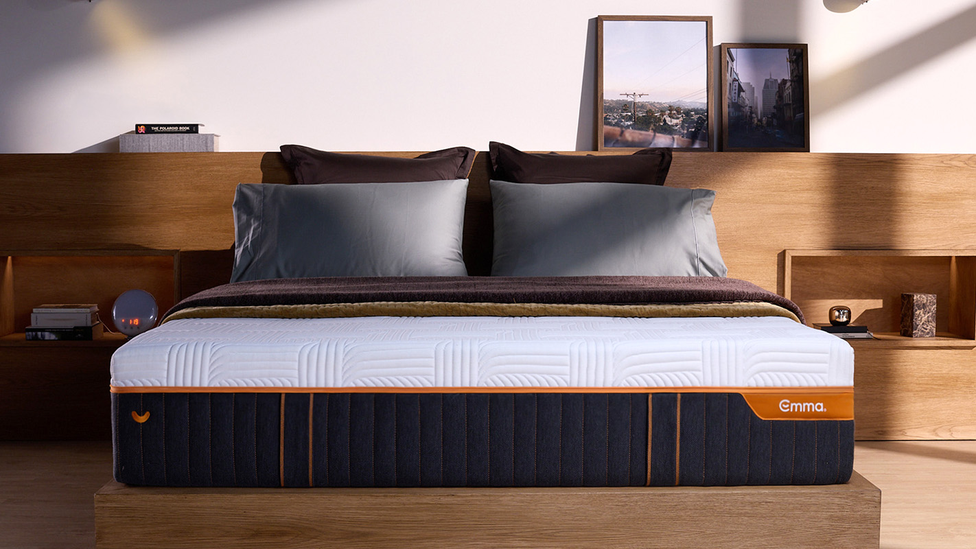 Front view of mattress in a room, showcasing it's height, dark base and white cover, with the orange Emma logo and zipper. 