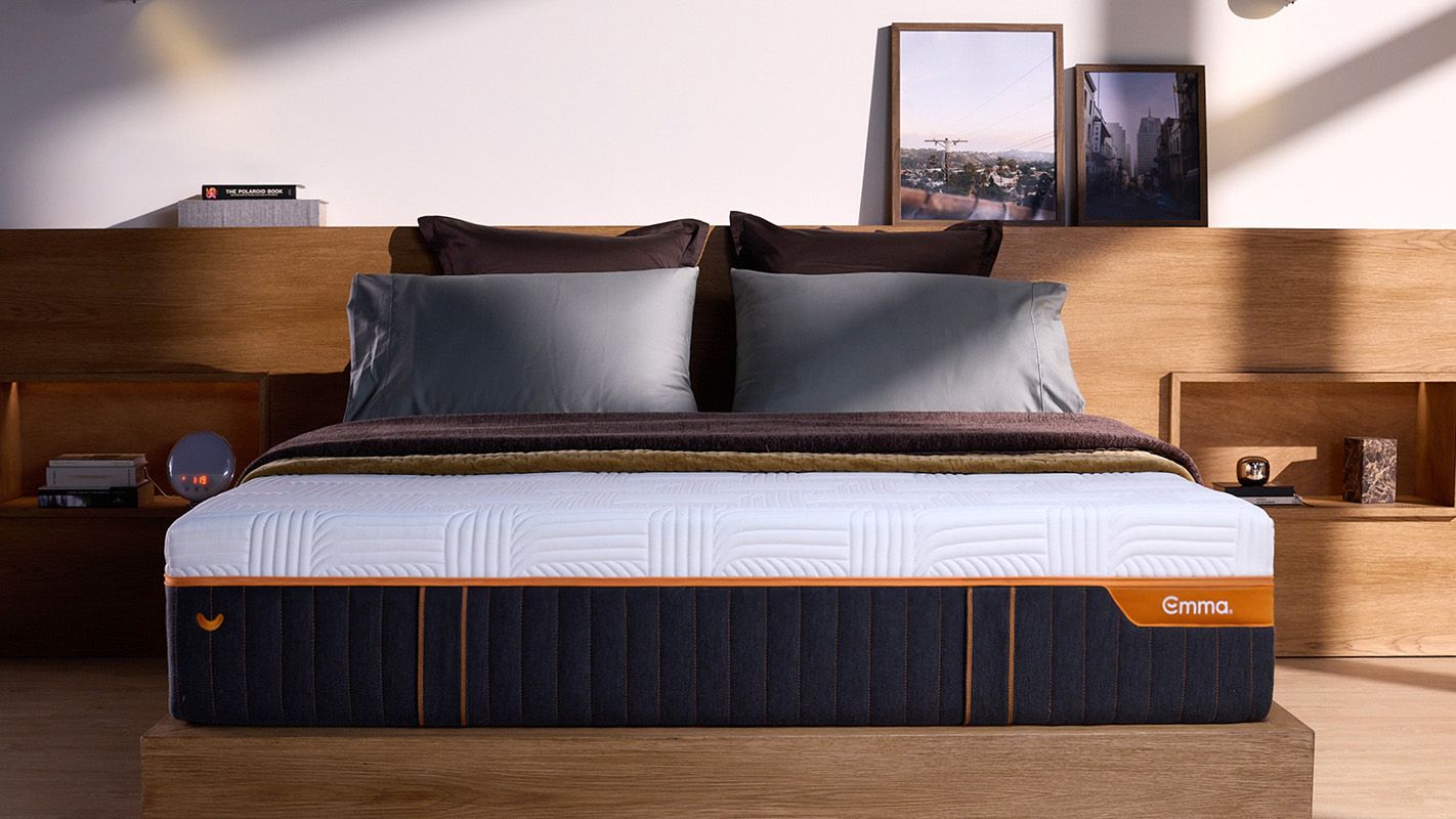Front view of mattress in a room, showcasing it's height, dark base and white cover, with the orange Emma logo and zipper.