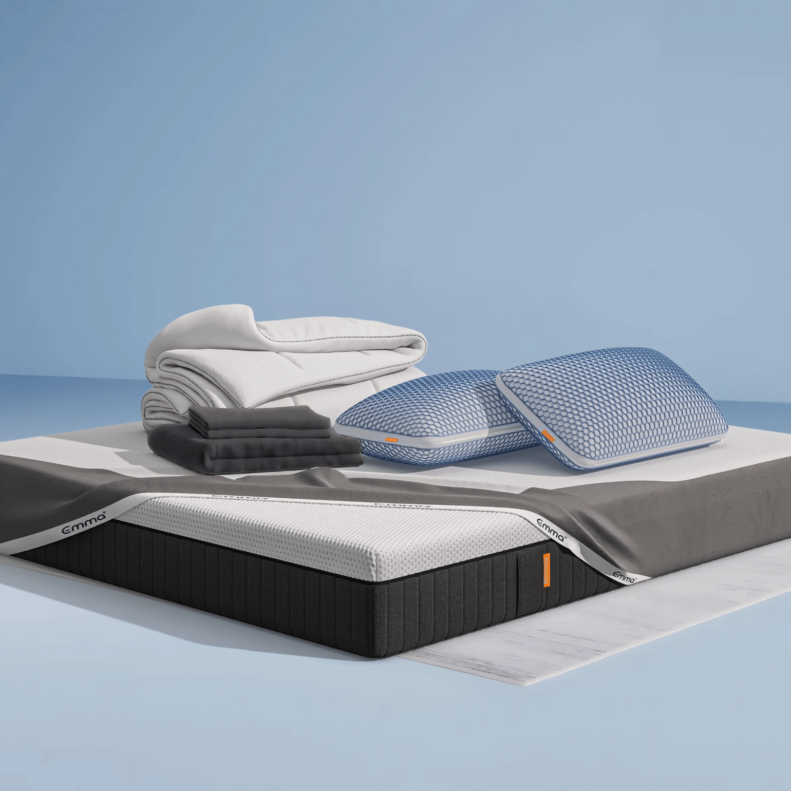 Ensemble de literie présenté dans une chambre moderne : matelas Hybride II ou Hybride Premium II avec base noire et surface blanche, partiellement recouvert d’un plaid gris foncé. Sur le matelas, deux oreillers Nuage Elite 40x70 cm bleus, une couette Nuage Tempérée blanche pliée, un protège-matelas, et une parure de lit en satin de coton blanc comprenant une housse de couette et des taies d’oreillers. Le tout est posé sur un tapis clair, avec une table d’appoint noire à pieds en bois et une décoration minimaliste.
