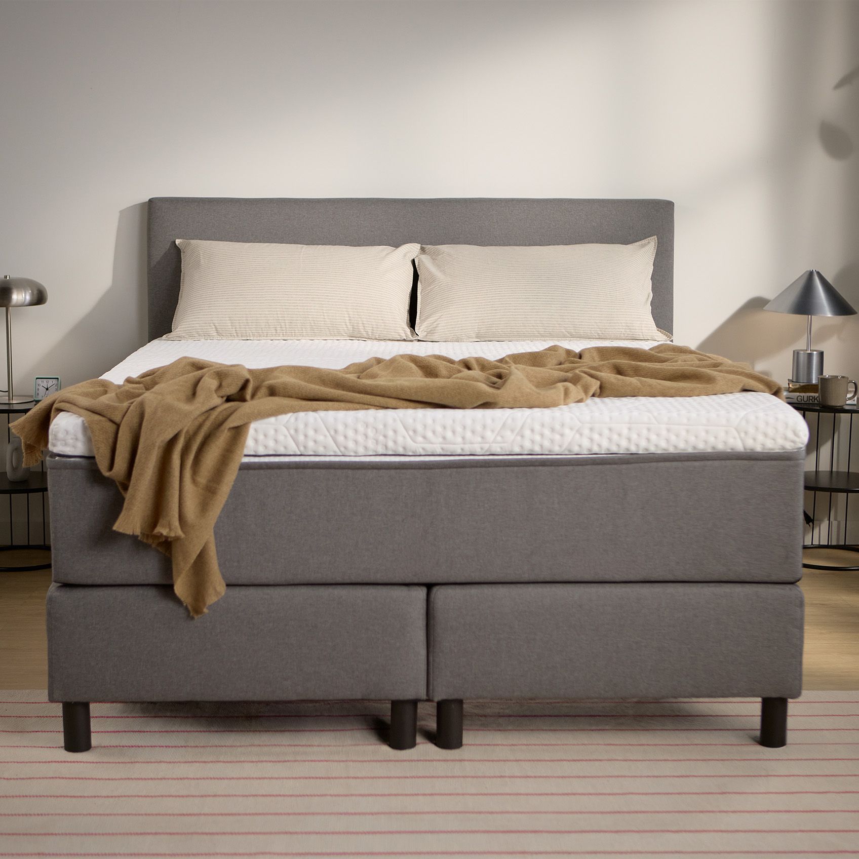 Dark grey upholstered box spring bed with headboard, visible central support legs, and bedding, shown from the front.