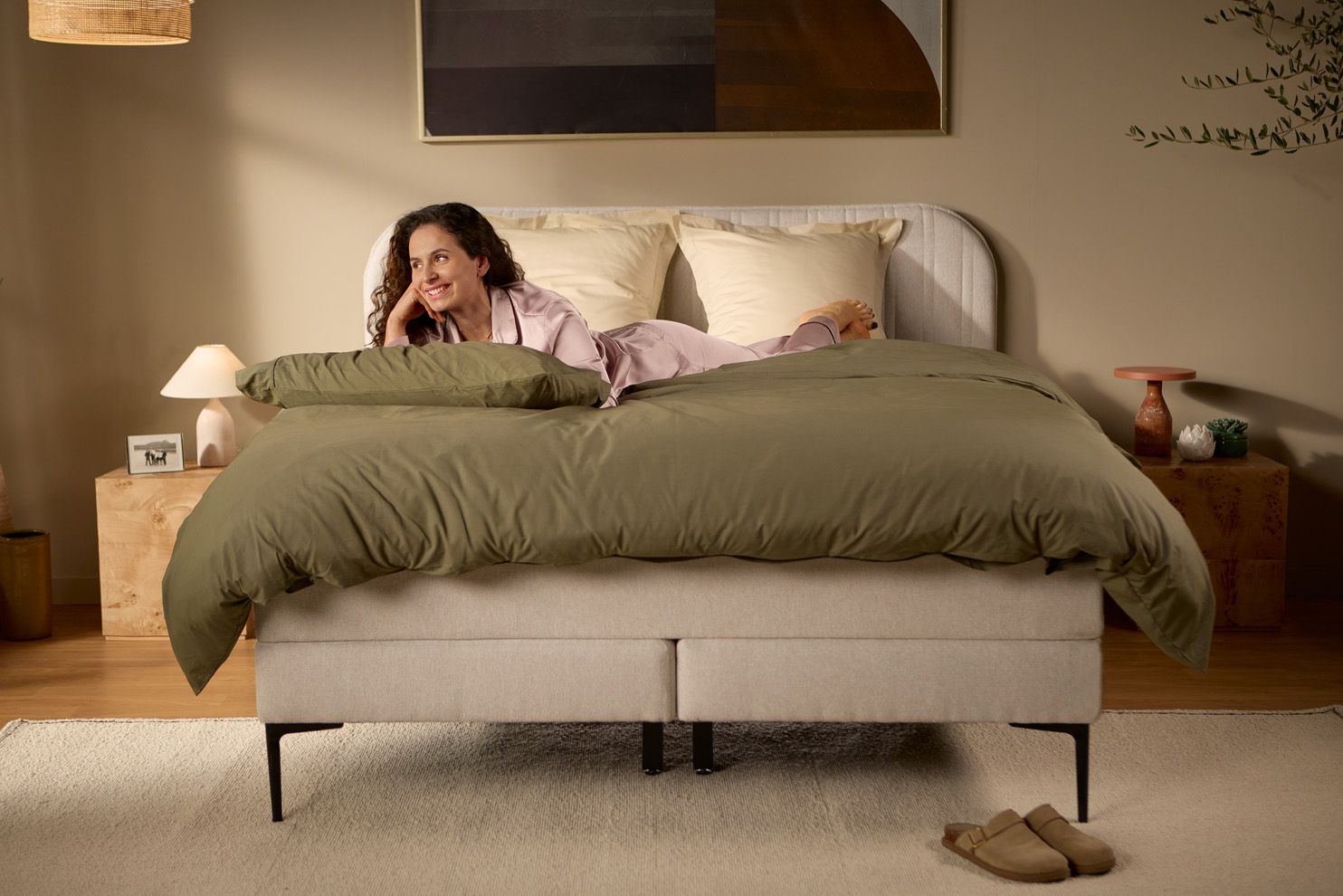 Iemand ligt op het bed met het beddengoed, waarbij de verhoogde boxspringbasis, het gestoffeerde hoofdbord en de zichtbare ondersteuningspoten goed te zien zijn.