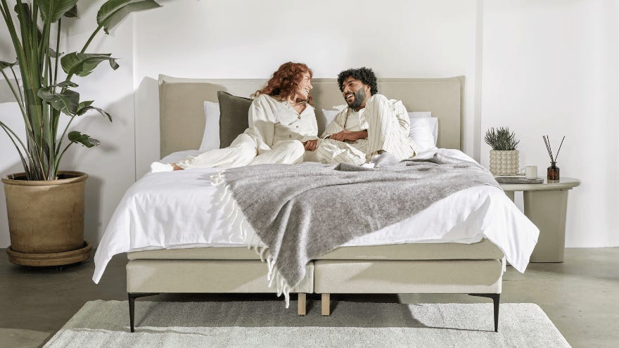 Twee mensen zitten ontspannen op een bed in een moderne, minimalistisch ingerichte slaapkamer met lichte kleuren, een grote kamerplant en een nachtkastje met een mok en decoratieve items.