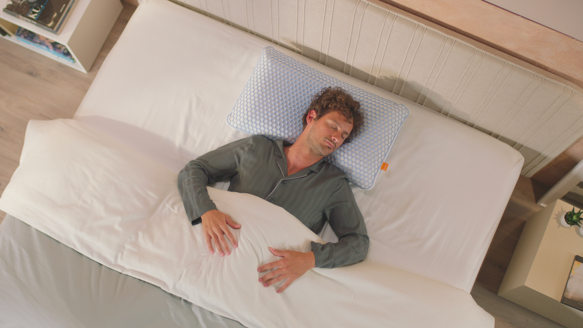 Persoon ligt op bed met hoofd op Emma AirGrid® Kussen met hexagonaal patroon en oranje label; witte deken, licht hoofdbord en nachtkastje met boeken en plant op achtergrond.