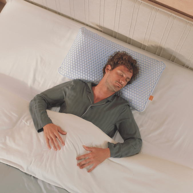 Persoon ligt op bed met hoofd op AirGrid® Adapt Kussen met hexagonaal patroon en oranje label; witte deken, licht hoofdbord en nachtkastje met boeken en plant op achtergrond.