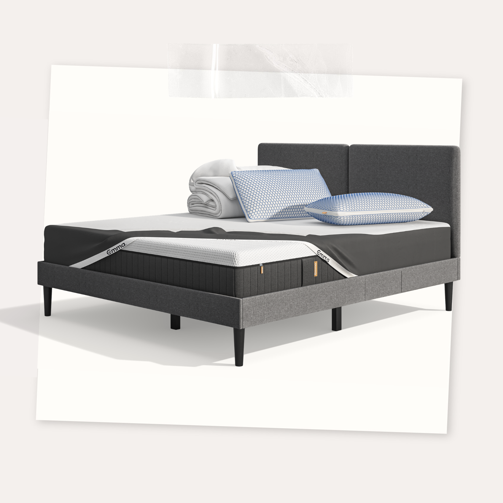 Lit moderne avec tête de lit grise et pieds noirs, recouvert de draps blancs. Deux oreillers blancs et une pile de linge plié sont posés sur le matelas. Arrière-plan beige avec une forme orange abstraite.