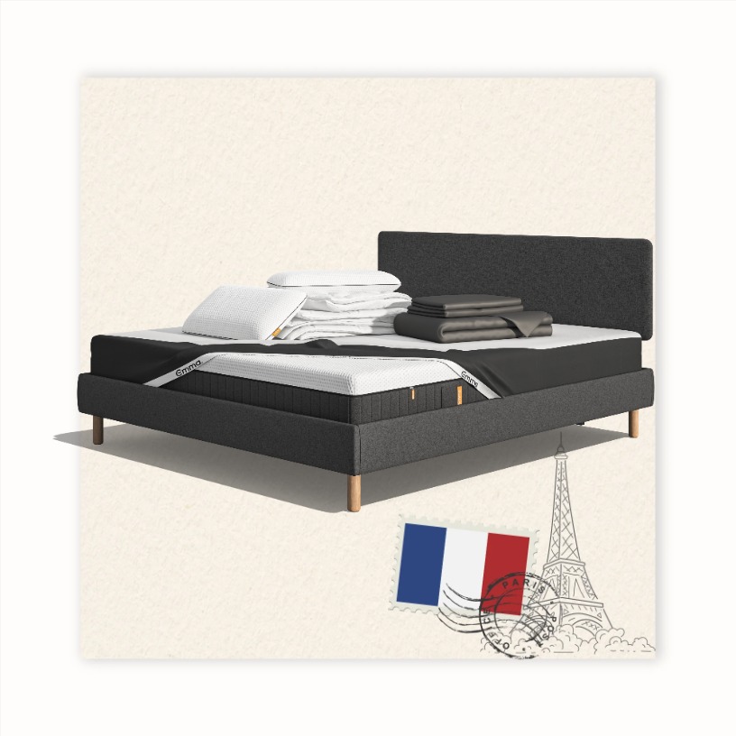 Lit moderne avec tête de lit grise et pieds noirs, recouvert de draps blancs. Deux oreillers blancs et une pile de linge plié sont posés sur le matelas. Arrière-plan beige avec une forme orange abstraite.
