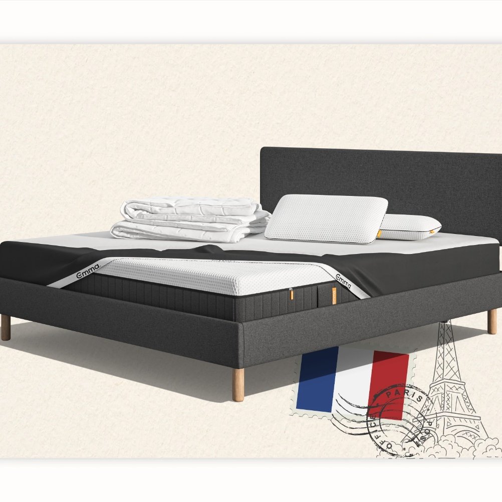 Lit moderne avec tête de lit grise et pieds noirs, recouvert de draps blancs. Deux oreillers blancs et une pile de linge plié sont posés sur le matelas. Arrière-plan beige avec une forme orange abstraite.
