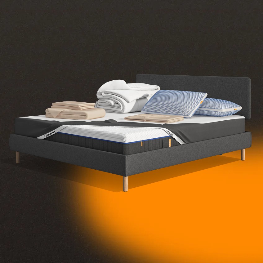 Lit moderne avec tête de lit grise et pieds noirs, recouvert de draps blancs. Deux oreillers blancs et une pile de linge plié sont posés sur le matelas. Arrière-plan beige avec une forme orange abstraite.