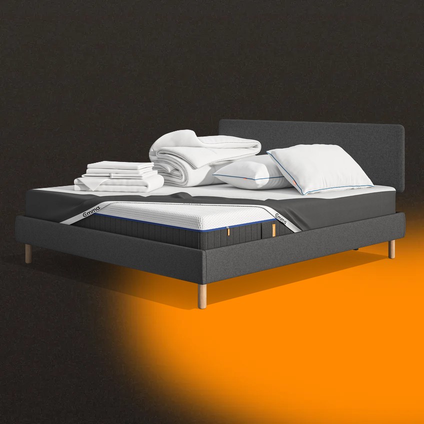 Lit moderne avec tête de lit grise et pieds noirs, recouvert de draps blancs. Deux oreillers blancs et une pile de linge plié sont posés sur le matelas. Arrière-plan beige avec une forme orange abstraite.