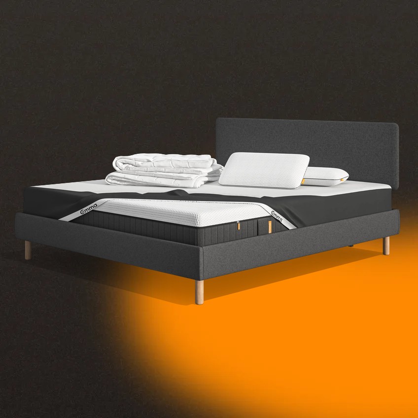 Lit moderne avec tête de lit grise et pieds noirs, recouvert de draps blancs. Deux oreillers blancs et une pile de linge plié sont posés sur le matelas. Arrière-plan beige avec une forme orange abstraite.