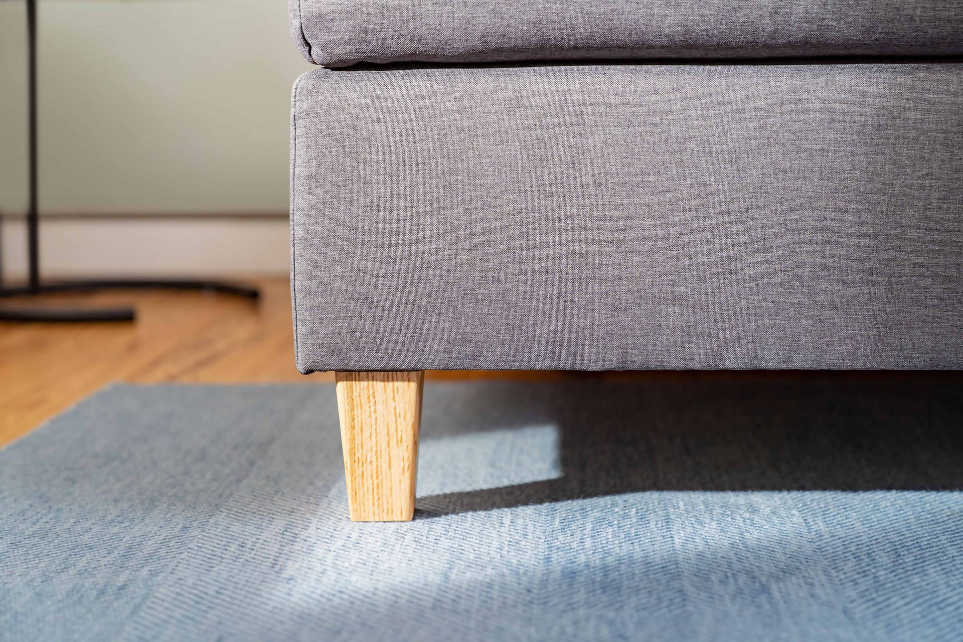 Detail van de grijze Emma Deluxe Boxspring met licht houten poot op een blauw textuurtapijt, geplaatst op een houten vloer in natuurlijk zonlicht.