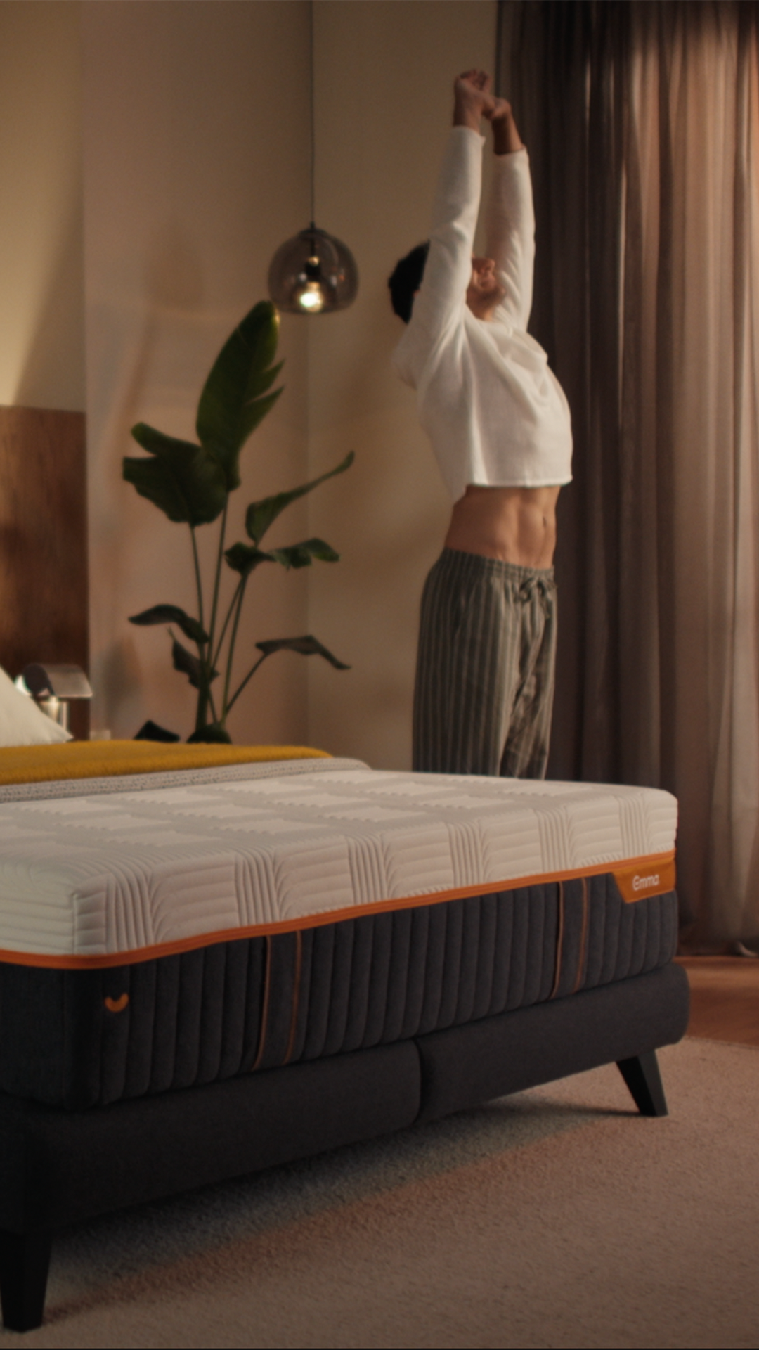Iemand die zich uitrekt na het ontwaken naast het Emma Performance-boxspringbed met een onderstel met oranje accenten in een stijlvol ingerichte slaapkamer.