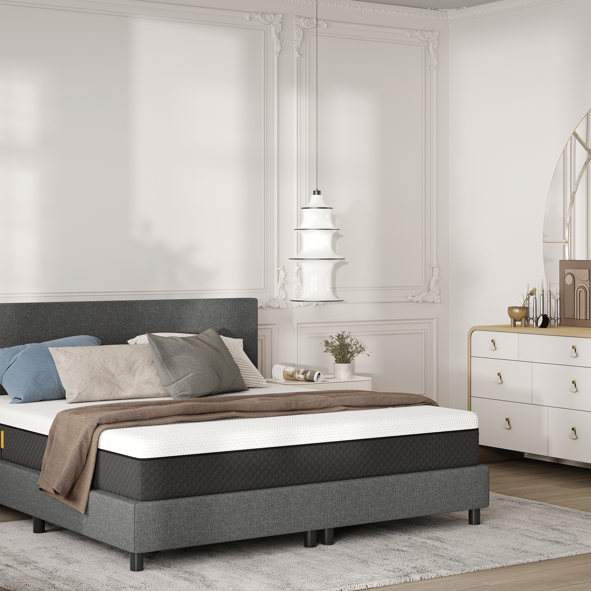 Lit avec tête de lit rembourrée gris foncé, matelas blanc à bordures sombres, et plusieurs oreillers dans des tons gris, beige et bleu. Couverture marron posée sur le lit. À droite, commode blanche et dorée avec objets décoratifs : livres, lampe et miroir rond doré. À gauche, fauteuil beige et table d’appoint avec vase de fleurs blanches. Murs ornés de moulures blanches, sol clair, lumière naturelle filtrée. Ambiance raffinée et minimaliste.