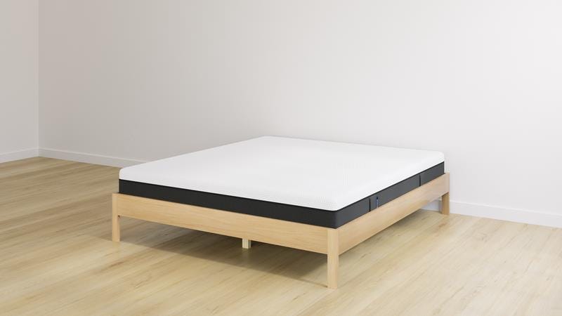 Emma houten bed met witte matras en zwarte onderzijde, geplaatst tegen een witte muur op een lichte houten vloer.