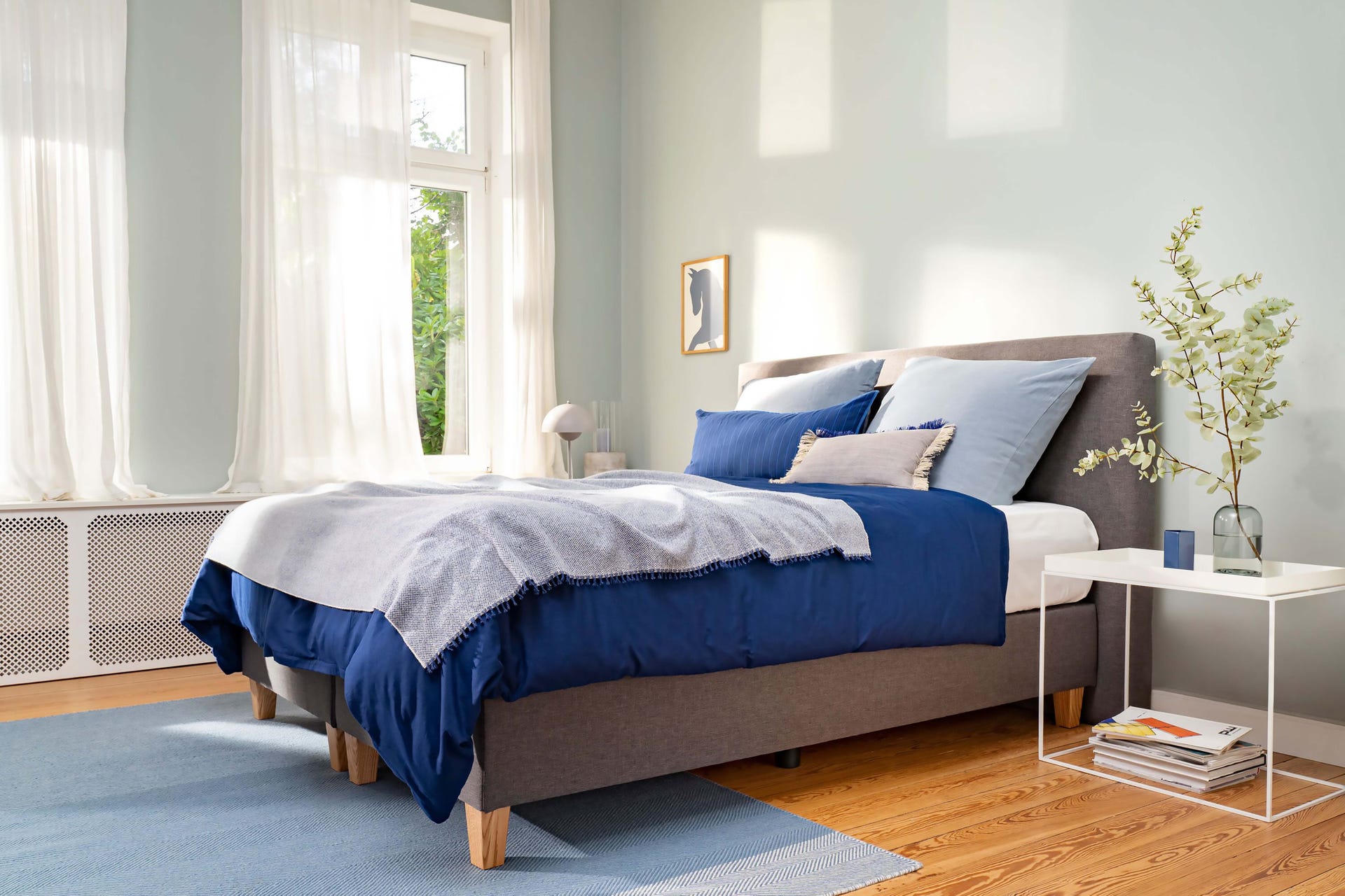 Emma Deluxe Boxspring in moderne slaapkamer: grijze gestoffeerde boxspring met houten poten, opgemaakt met blauw-witte beddengoed, naast een wit nachtkastje met decoratieve vaas, boeken en lamp, in een lichte ruimte met houten vloer en grote ramen.