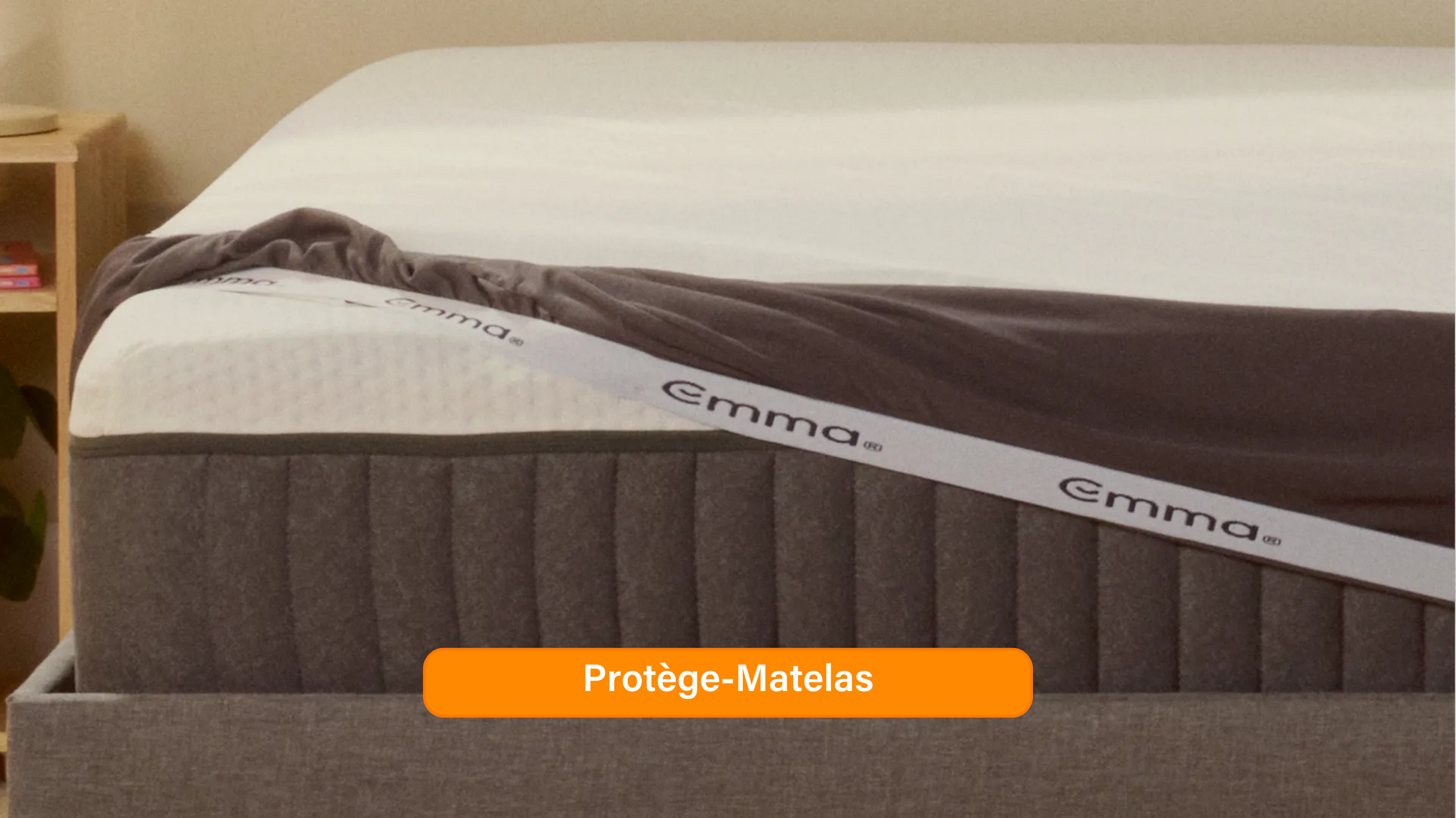 Gros plan sur le Protège-Matelas Emma partiellement retiré, révélant la surface blanche texturée et perforée du matelas en dessous. Le protège-matelas est gris et blanc, avec le logo Emma visible sur le côté. Une étiquette orange en bas de l’image indique "Protège-Matelas".