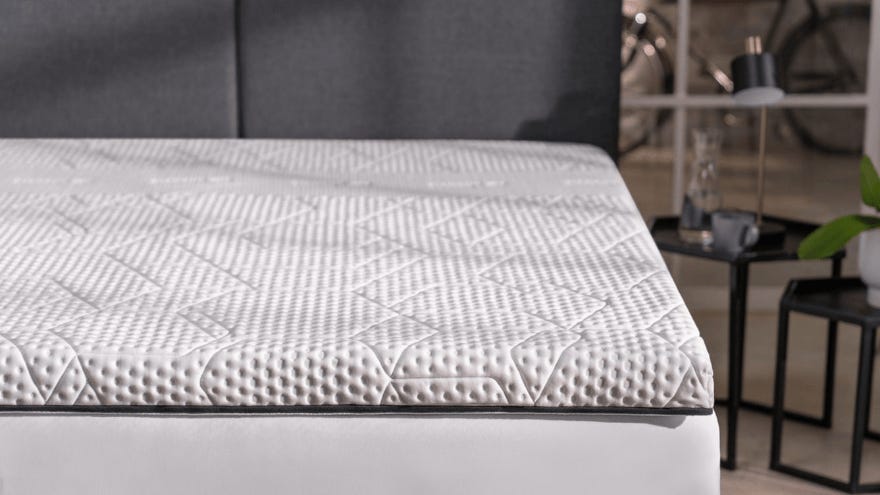 Close-up van een bed met een matras-topper met textuur. Het oppervlak toont geometrische patronen zoals stippen, lijnen, driehoeken en diamantvormen. Op de achtergrond zijn een grijze hoofdbord, een bijzettafel met een glas water, een kopje en een plantje zichtbaar. Verderop staan een fiets en een boekenkast.