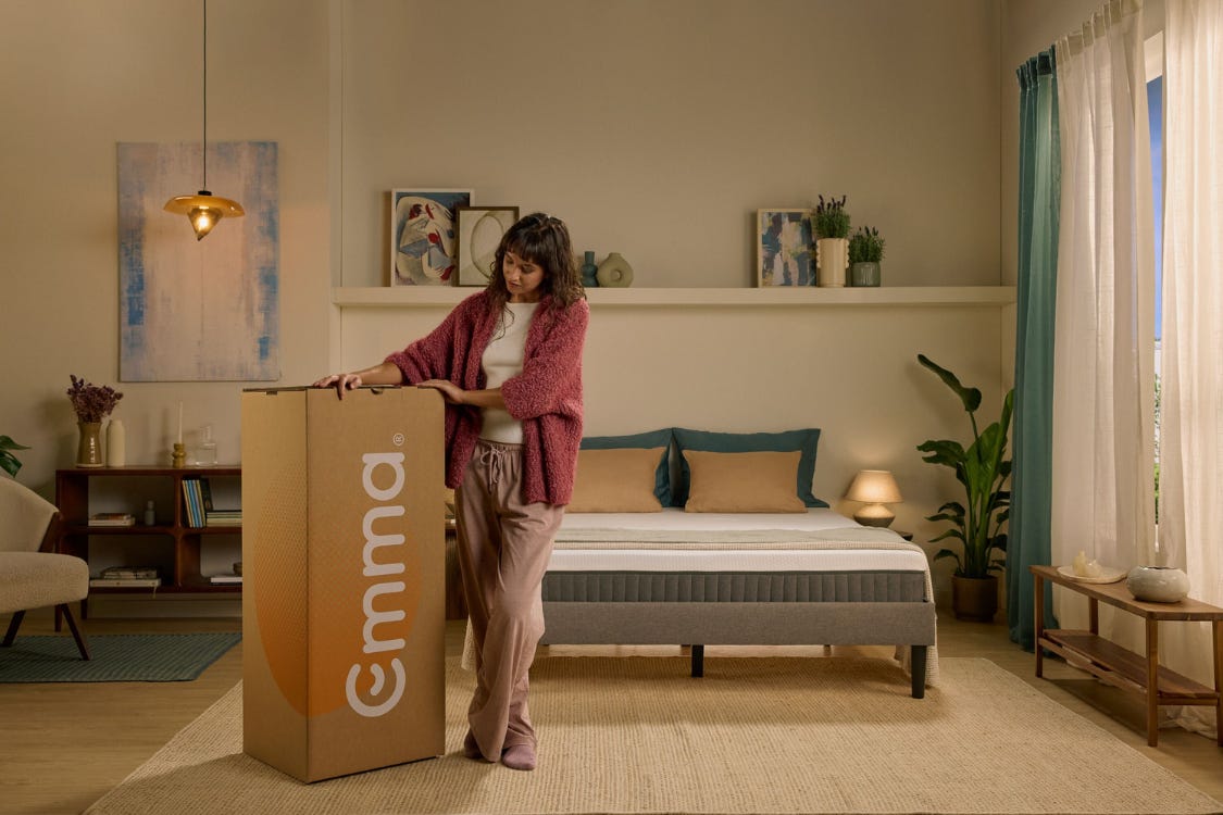 A girl standing next to an Emma box, in front of an Emma bed and mattress.