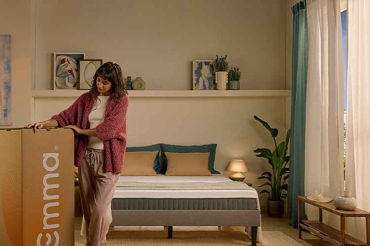 Une femme est assise sur un lit dans une chambre lumineuse et décorée avec goût, enveloppée dans une couette blanche. Elle regarde vers le haut avec une expression paisible. La pièce contient un fauteuil orange, des plantes vertes, des cadres au mur et des objets décoratifs, créant une ambiance chaleureuse et minimaliste.