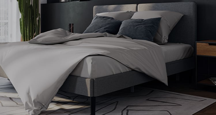 Quarto moderno com cama de estrutura e cabeceira estofadas em cinza, da marca Emma, coberta por roupa de cama em tons de cinza claro e almofadas decorativas escuras. Ao lado da cama, há um criado-mudo de madeira com prateleira e gaveta. À esquerda, uma planta alta em vaso próximo à janela com cortinas translúcidas que deixam entrar luz natural.