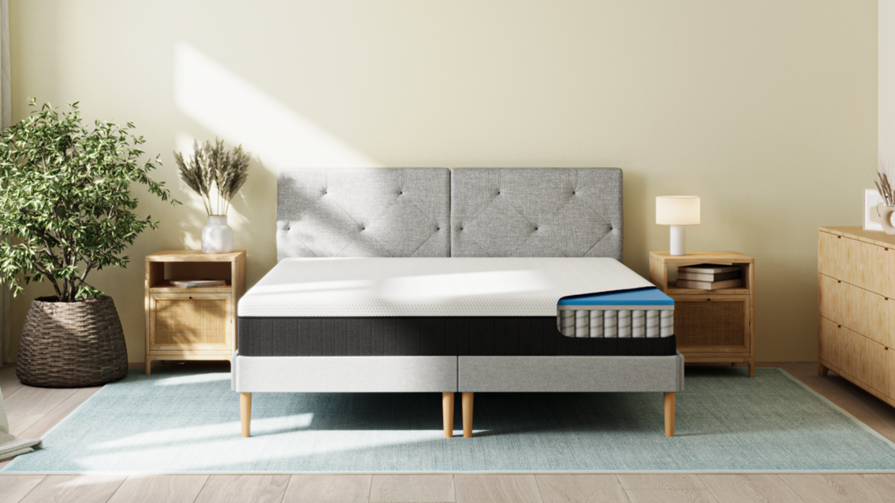 Chambre moderne avec lit équipé du Matelas Original II, dont une section coupée révèle les couches internes : mousse supérieure blanche, couche bleue de transition, et base à ressorts ensachés. Le lit a une tête de lit gris clair capitonnée et repose sur des pieds en bois. Deux tables de chevet en bois avec tiroirs en rotin encadrent le lit, chacune avec une lampe et des objets décoratifs. À gauche, une grande plante en pot ; à droite, une commode en bois. Le sol est en bois clair avec un tapis bleu clair, et la lumière naturelle entre par la gauche.