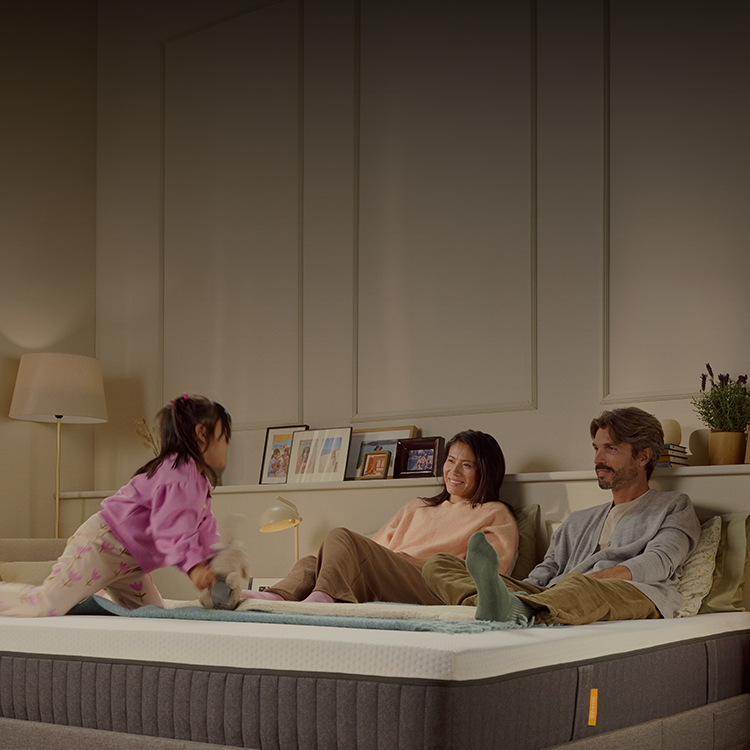 Une femme allongée sur le côté dort paisiblement sur un matelas, la tête posée sur ses mains. La scène se déroule dans une chambre lumineuse avec une fenêtre en arrière-plan et une plante verte à côté du lit, créant une atmosphère calme et reposante.