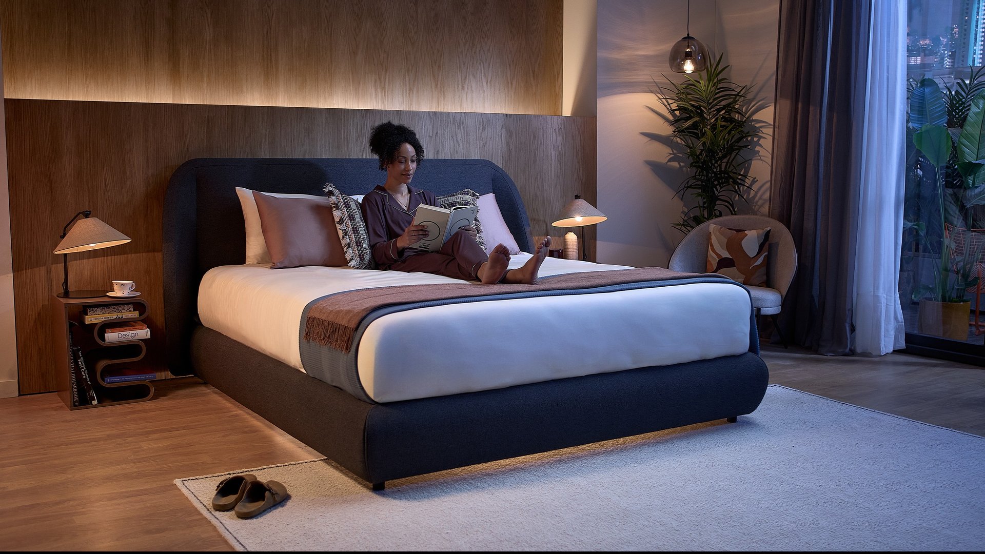 Deux personnes allongées face à face dans un lit, sous une couette bleu clair à motifs, dans une chambre paisible avec éclairage doux et table de nuit garnie d’objets du quotidien.
