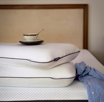 Emma Diamond Kussen met witte hoes en zwarte rits, gestapeld op bed met beige hoofdbord—kopje en schotel bovenop kussen benadrukken stabiliteit en comfort.