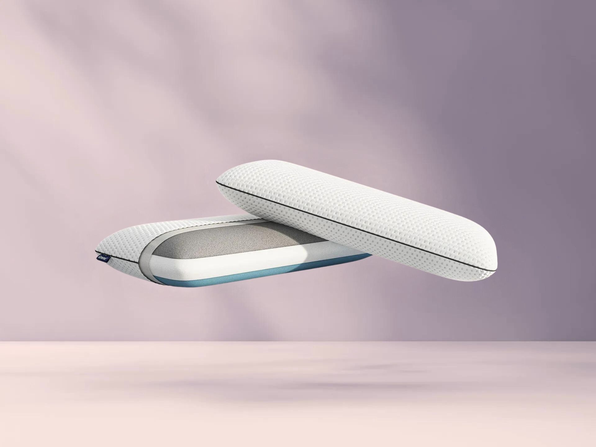 Twee witte Original Cooling Adapt Kussens met blauwe bies zweven tegen zachte paarse achtergrond; één kussen toont grijze binnenlaag via geopende rits, benadrukt luxe en aanpasbaarheid van de set.