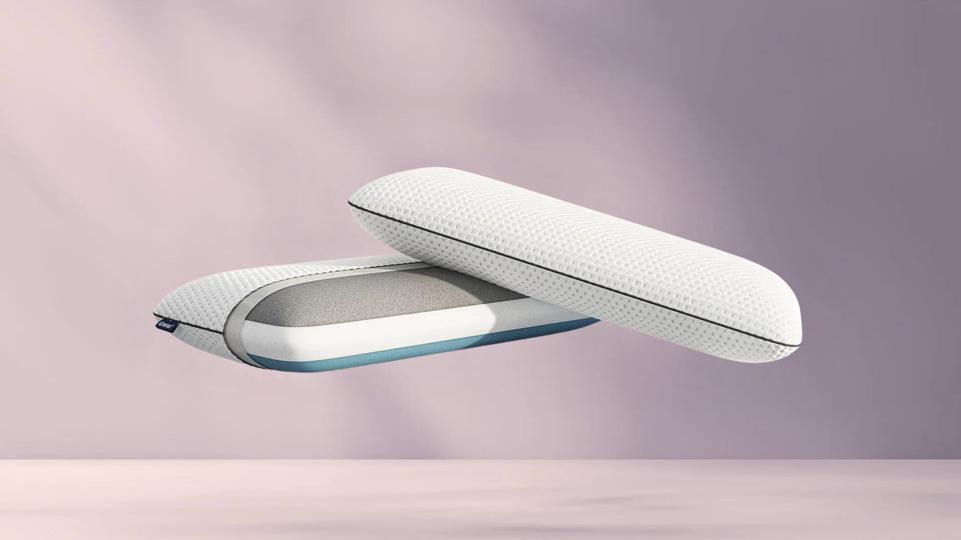 Twee witte Original Cooling Adapt Kussens met blauwe bies zweven tegen zachte paarse achtergrond; één kussen toont grijze binnenlaag via geopende rits, benadrukt luxe en aanpasbaarheid van de set.