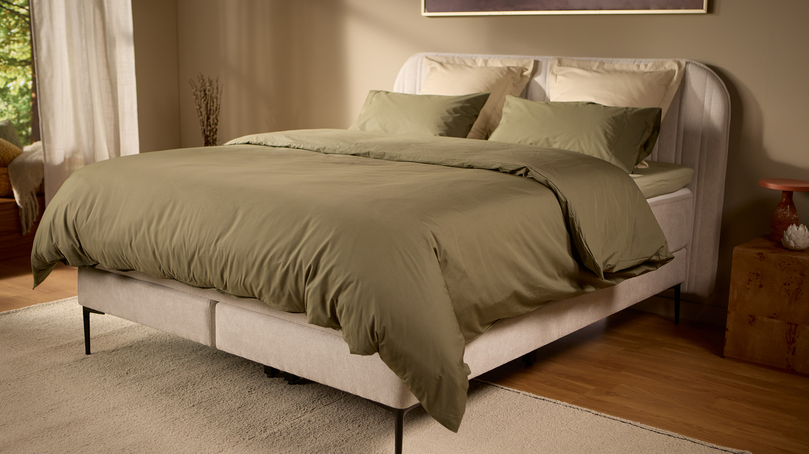 Emma Boxspring Bed in beige with curved channelled headboard styled with olive green bedding in a warm bedroom.
