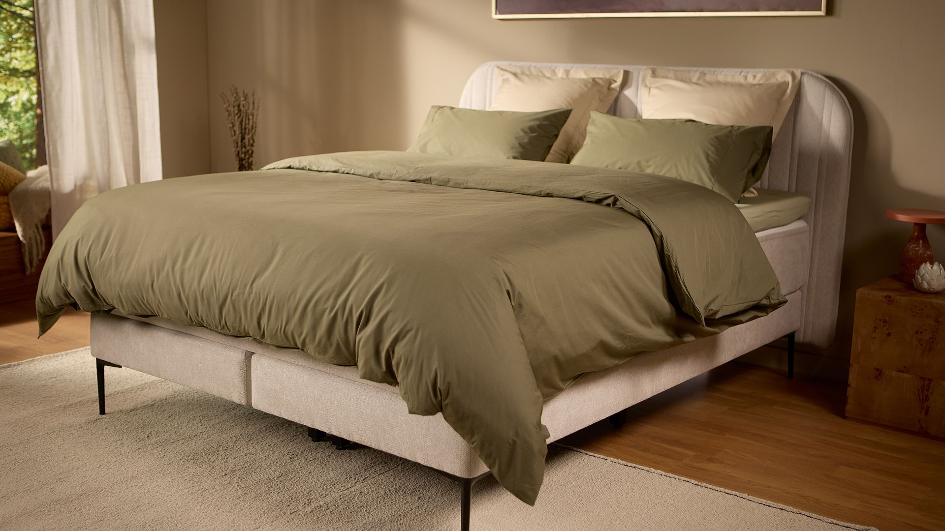 Emma Boxspring Bed in beige with curved channelled headboard styled with olive green bedding in a warm bedroom.
