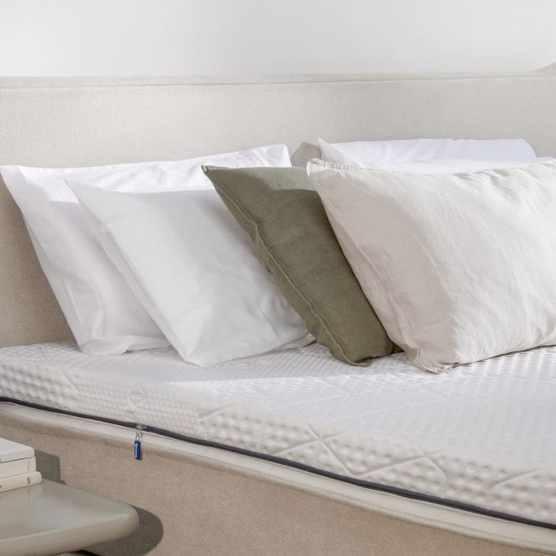 Letto rifatto con testiera beige, quattro cuscini e topper bianco testurizzato; comodino con libri, tazza e pianta decorativa
