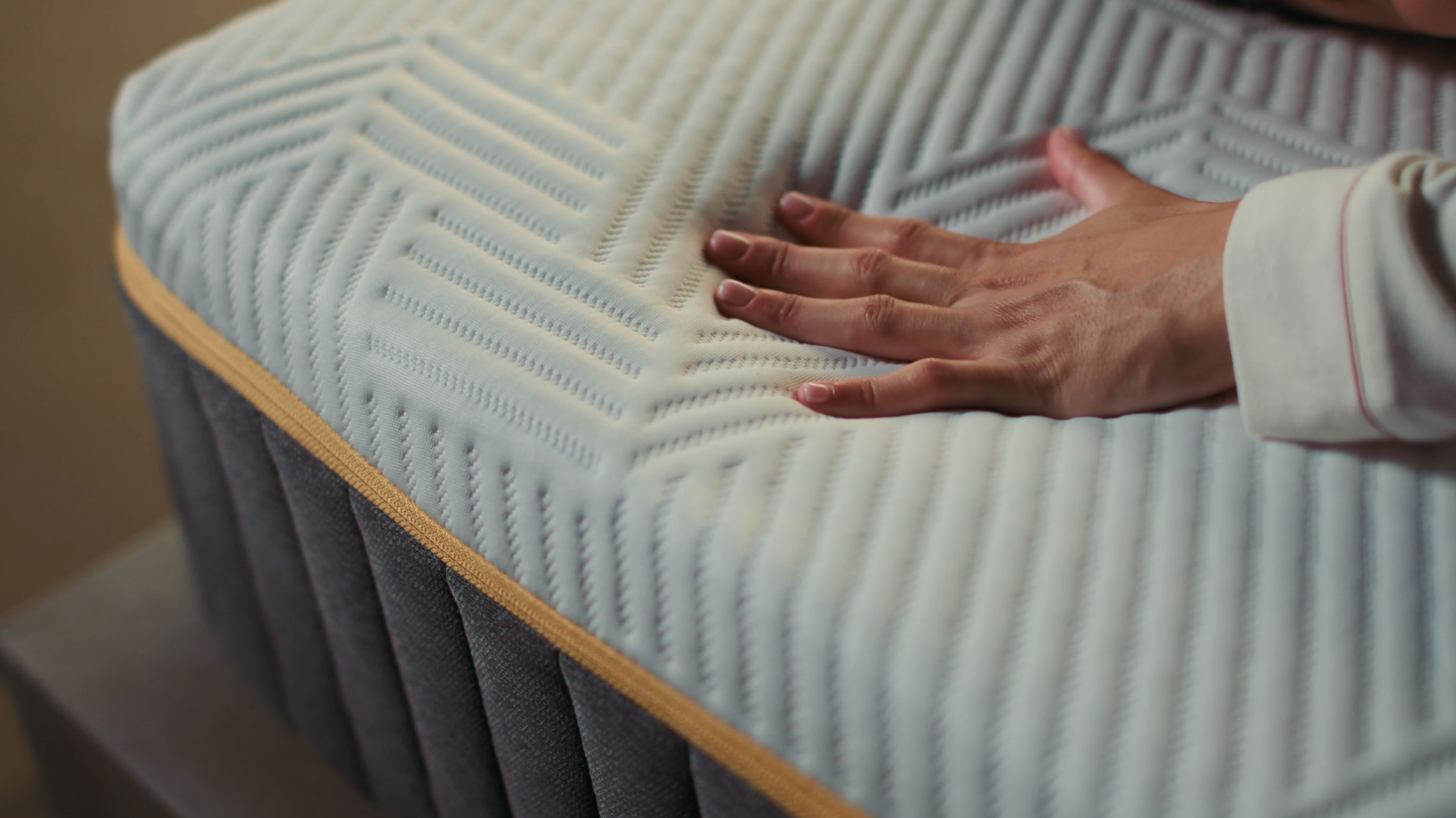 Hand pressing into the corner of an Emma Original Pro mattress, showing the textured cover and gold trim in close-up detail.
