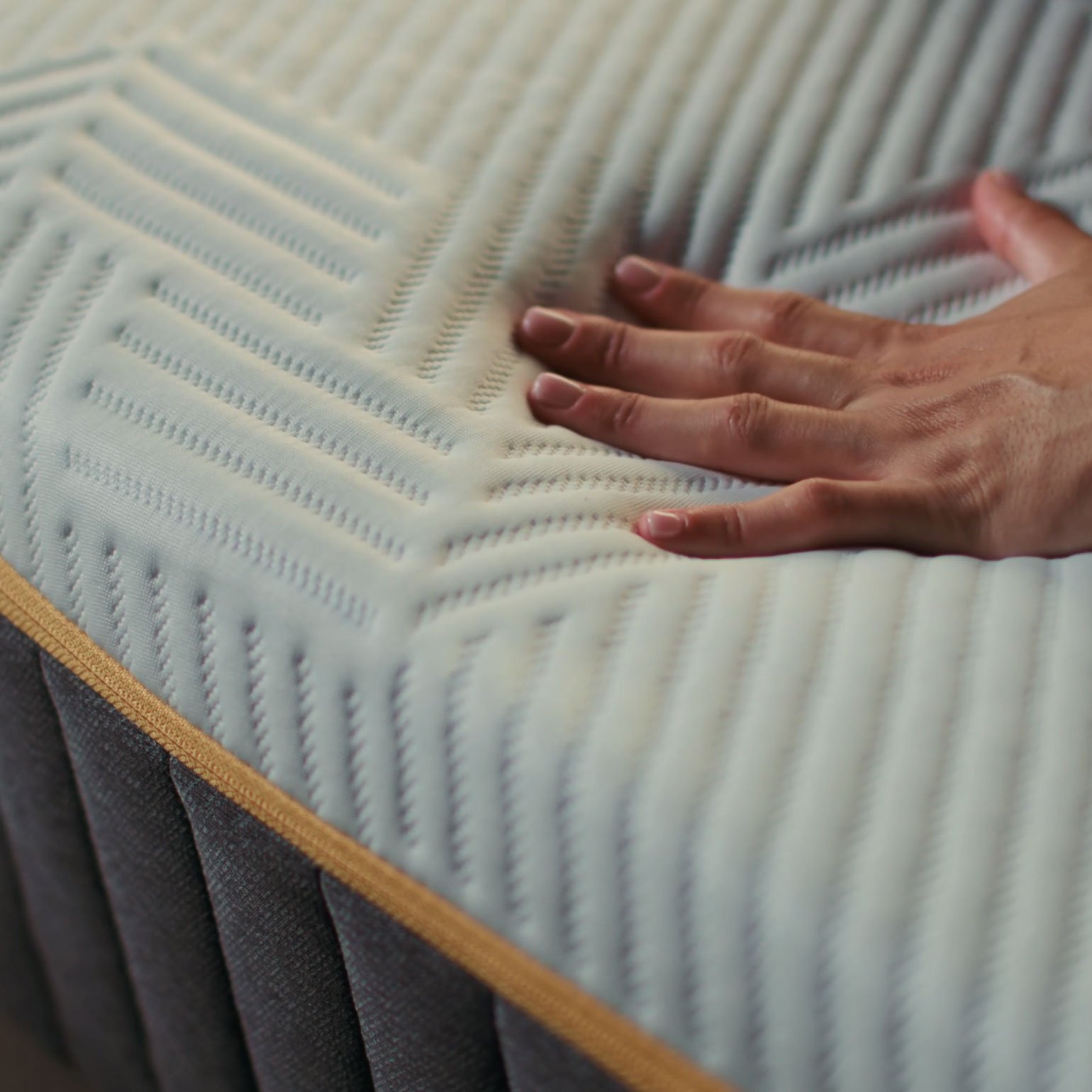 Hand pressing into the corner of an Emma Original Pro mattress, showing the textured cover and gold trim in close-up detail.
