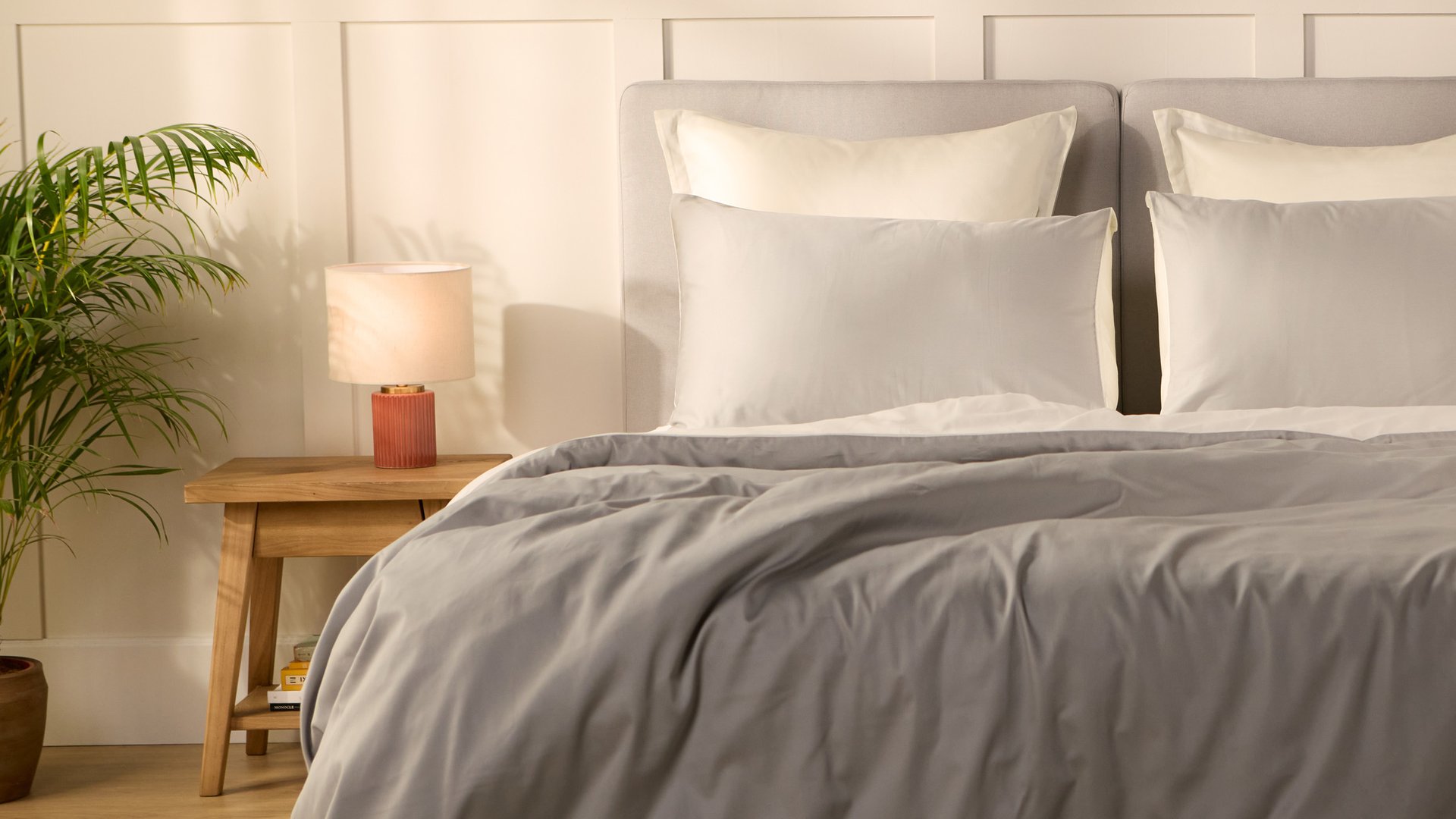 Slaapkamer met een opgemaakt bed in lichtgrijs beddengoed, met een houten nachtkastje met lamp en een groene plant ernaast.