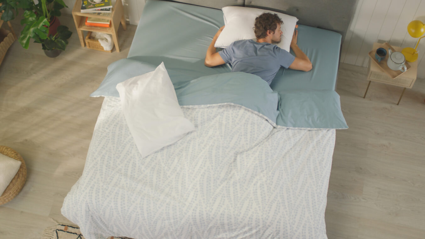 Persoon ligt voorover op bed met lichtblauwe lakens en wit Emma Premium Wolkenkussen; nachtkastje met gele lamp, kopje en boek; plank met boeken en plant op achtergrond.