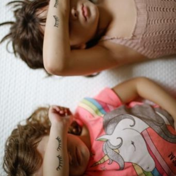 Duas crianças deitadas sobre o Colchão Emma Original. Têm o braço direito com pestanas desenhadas, tapando seus olhos. 