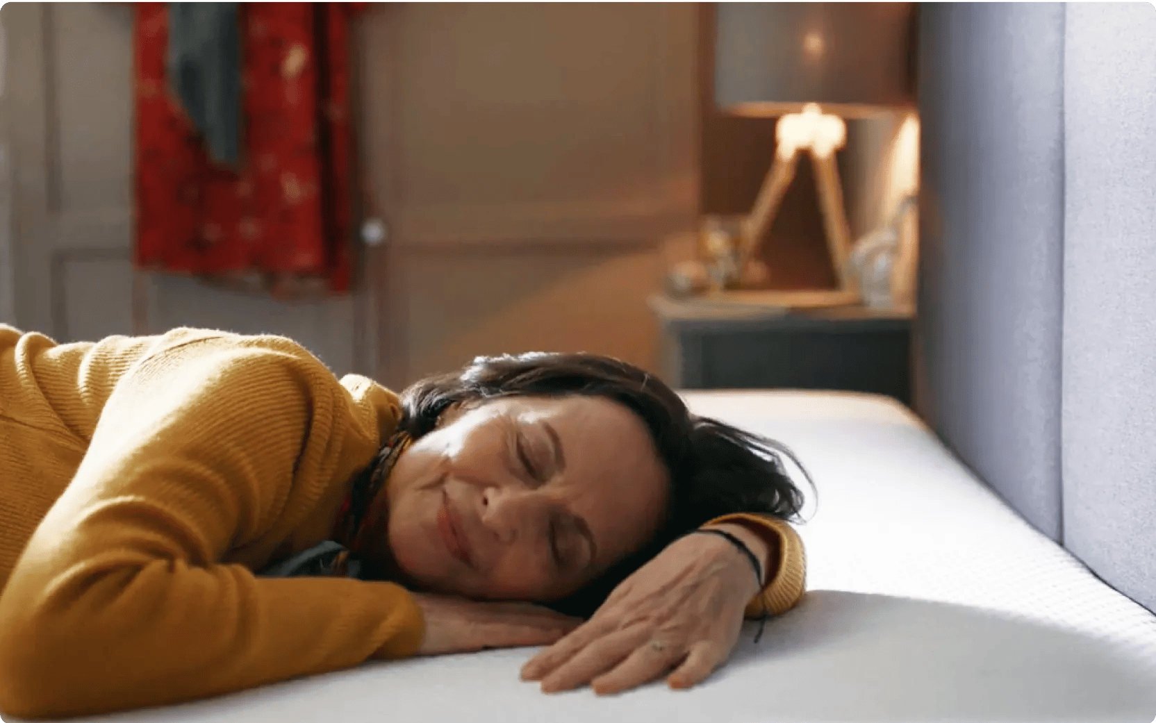 Persoon ligt ontspannen op een Emma-matras, gekleed in een okergele trui. Op de achtergrond een nachtkastje met lamp en decoratie, en een rood kledingstuk aan de deur. De setting straalt comfort en huiselijkheid uit.