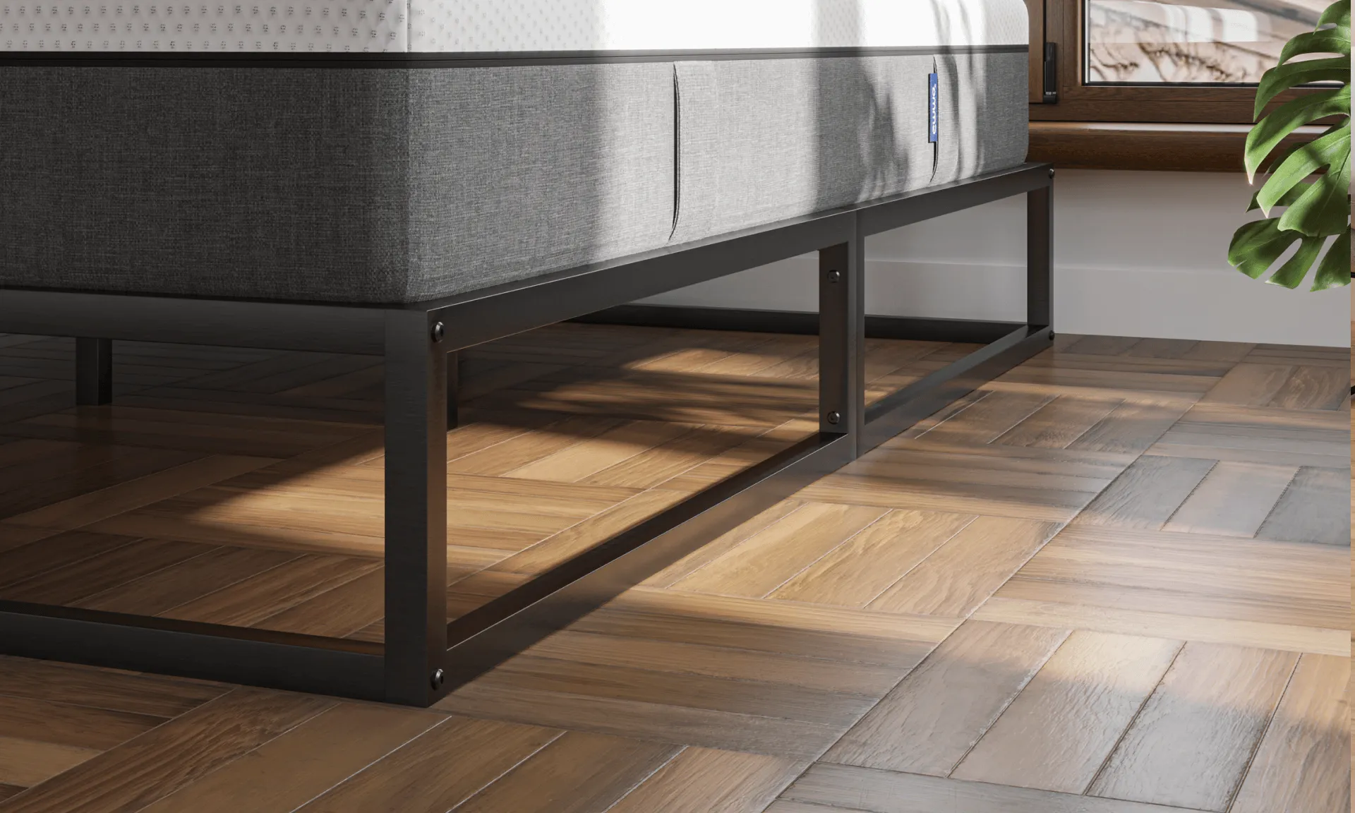 Minimalistisch metalen bedframe in zwarte afwerking, geplaatst op visgraat houten vloer; bed is voorzien van wit matras met grijze zijpanelen en staat voor raam met uitzicht op dakpannen en een groene plant in de hoek.