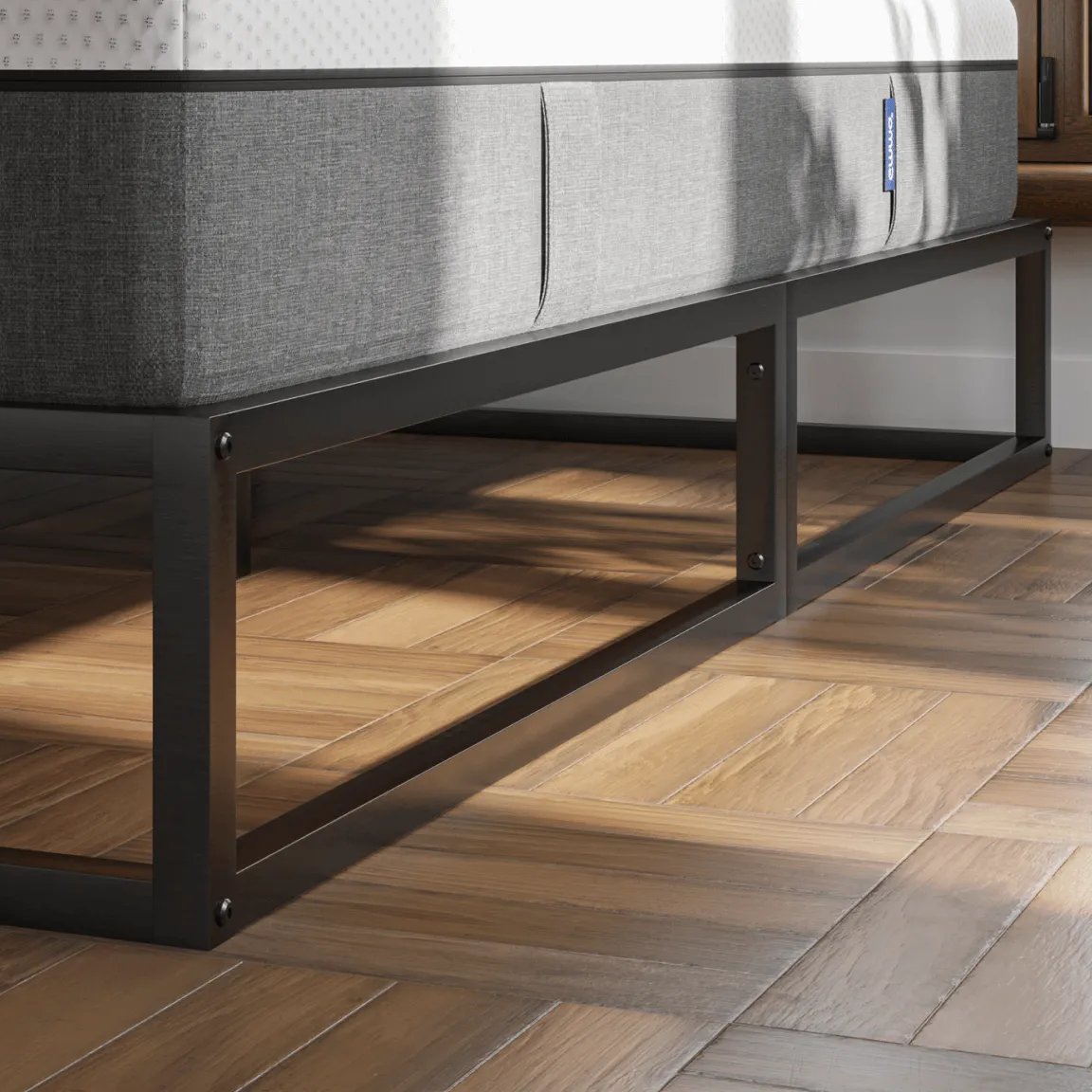 Minimalistisch metalen bedframe in zwarte afwerking, geplaatst op visgraat houten vloer; bed is voorzien van wit matras met grijze zijpanelen en staat voor raam met uitzicht op dakpannen en een groene plant in de hoek.
