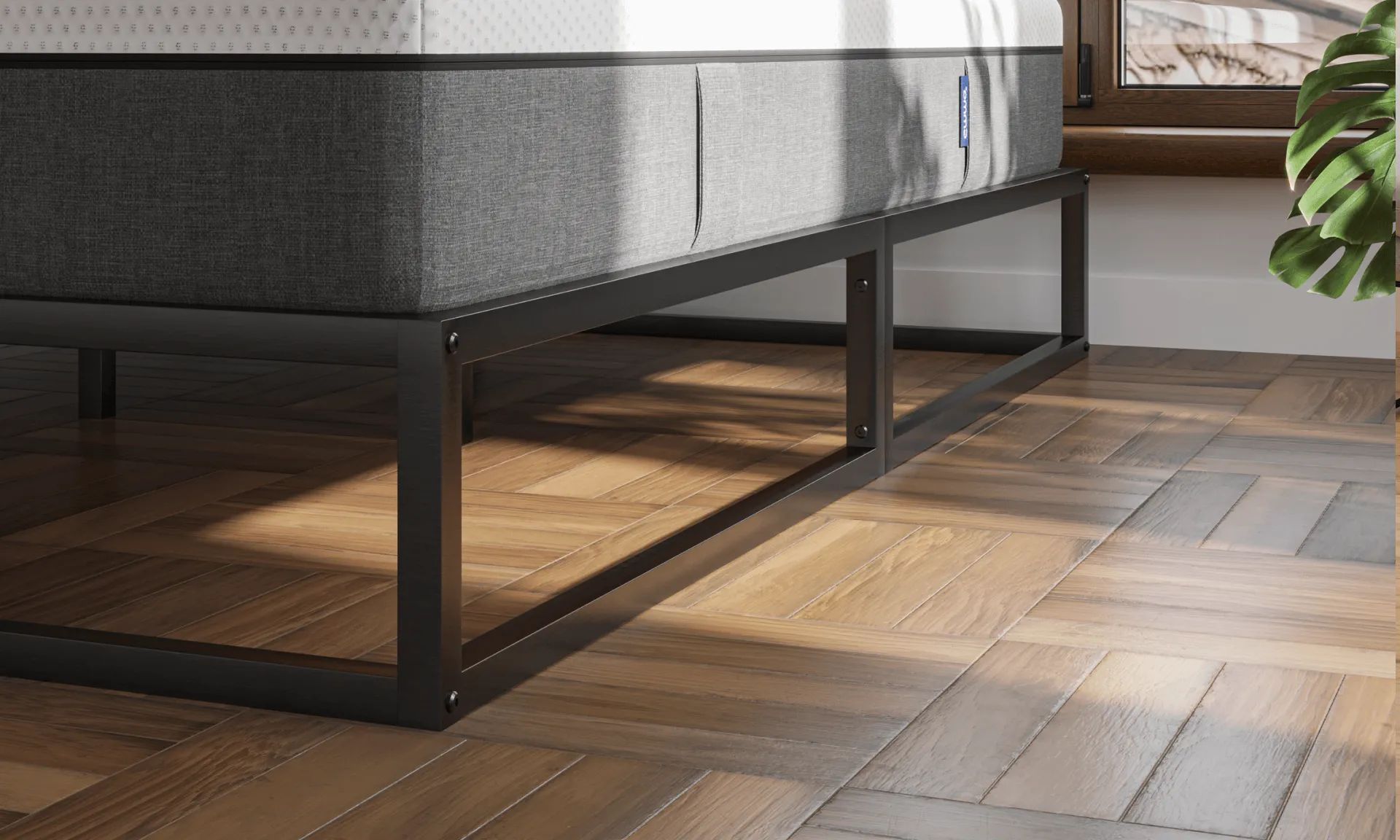 Minimalistisch metalen bedframe in zwarte afwerking, geplaatst op visgraat houten vloer; bed is voorzien van wit matras met grijze zijpanelen en staat voor raam met uitzicht op dakpannen en een groene plant in de hoek.