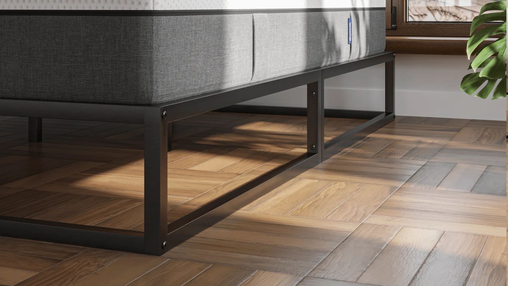 Minimalistisch metalen bedframe in zwarte afwerking, geplaatst op visgraat houten vloer; bed is voorzien van wit matras met grijze zijpanelen en staat voor raam met uitzicht op dakpannen en een groene plant in de hoek.