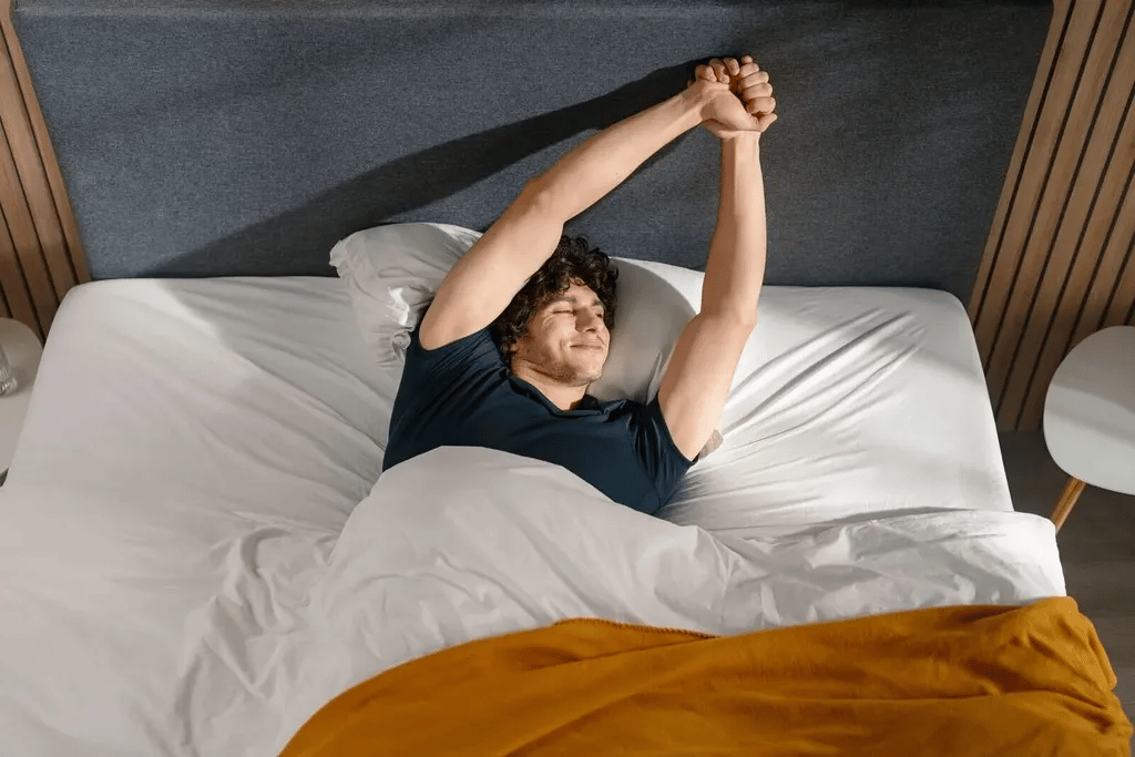 Persoon ligt in bed en strekt beide armen boven het hoofd, onder een witte deken met een okergele sprei aan het voeteneinde.